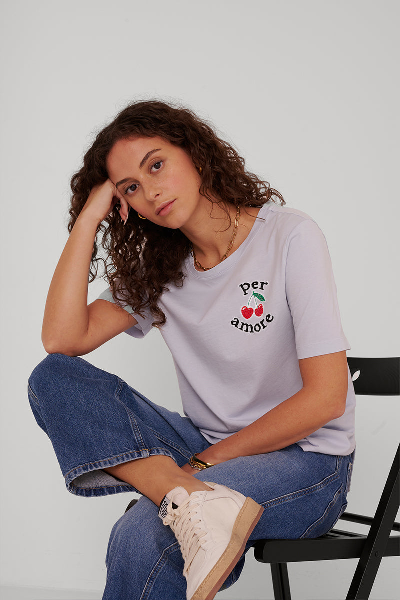 Eine Frau mit lockigem braunem Haar sitzt auf einem schwarzen Stuhl, den Ellbogen auf dem Knie, den Kopf auf die Hand gestützt. Sie trägt ein helllavendelfarbenes Princess Goes Hollywood-T-Shirt mit Kirschstickerei, blaue Jeans und rosa Turnschuhe vor einem schlichten weißen Hintergrund.