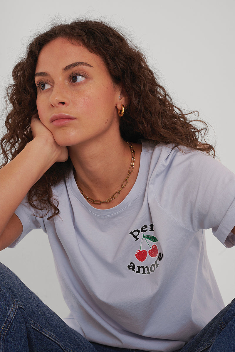 Eine junge Frau mit lockigen braunen Haaren trägt ein hellviolettes "per amore" Baumwolle T-Shirt mit Kirschstickerei von Princess Goes Hollywood, gepaart mit blauen Jeans, goldenen Ohrringen und einer Kettenhalskette vor einem neutralen Hintergrund.