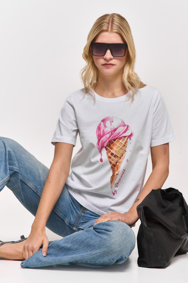 Eine Frau mit lockeren blonden Wellen sitzt auf dem Boden vor einem weißen Hintergrund, trägt eine Princess Goes Hollywood-Sonnenbrille, blaue Jeans, schwarze Sandalen, ein rosa T-Shirt mit Eis-Print Baumwolle und hat eine große schwarze Tasche neben sich.