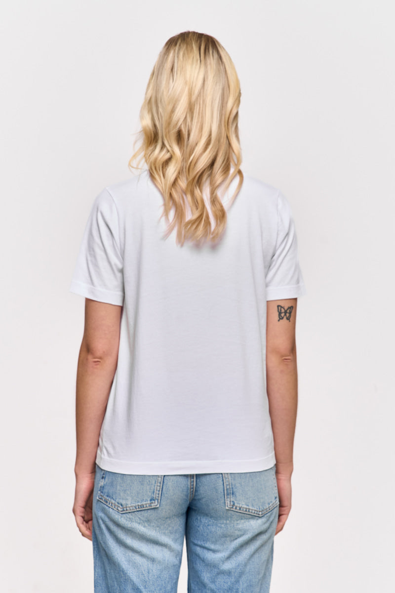 Eine Person mit gewelltem blondem Haar ist von hinten zu sehen. Sie trägt ein schlichtes weißes Baumwolle T-Shirt von Princess Goes Hollywood und hellblaue Jeans. Auf ihrem linken Oberarm ist ein kleines Schmetterlingstattoo auf weißem Hintergrund zu sehen.