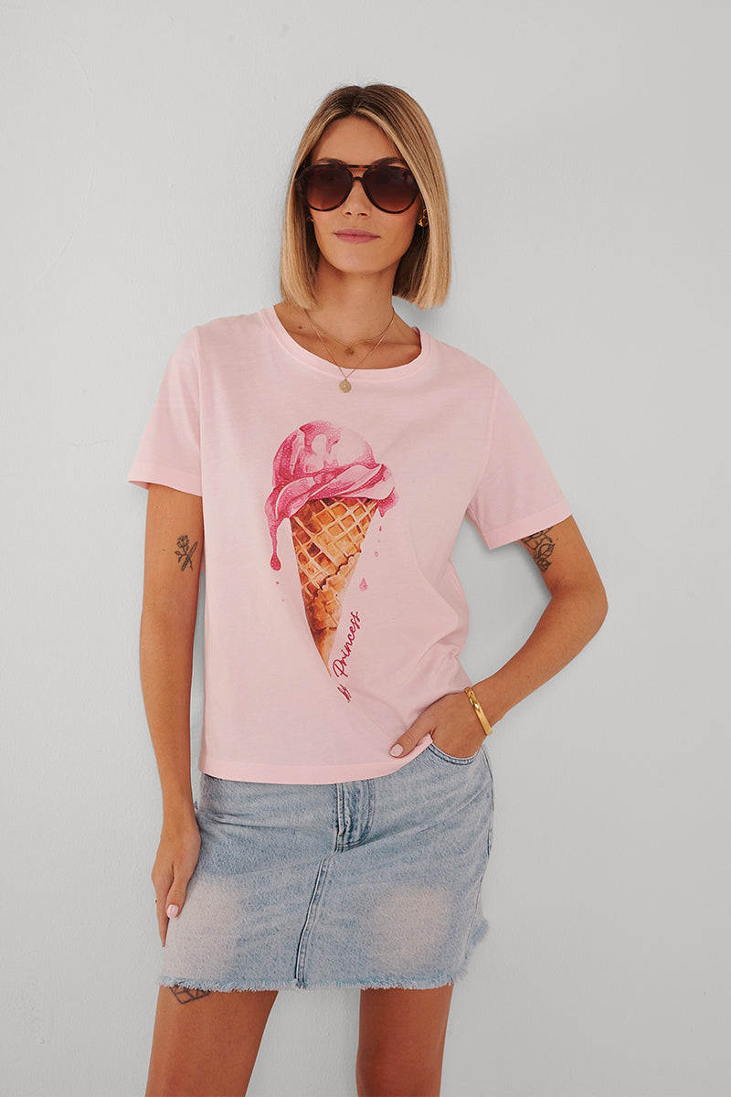 Eine Frau mit glattem blondem Haar trägt eine große Sonnenbrille, Goldschmuck und ein hellrosa Princess Goes Hollywood-T-Shirt mit einer Grafik von schmelzendem Eis. Ihr Jeansrock hat Gürtelschlaufen und einen ausgefransten Saum, während sie vor einer weißen Wand steht.