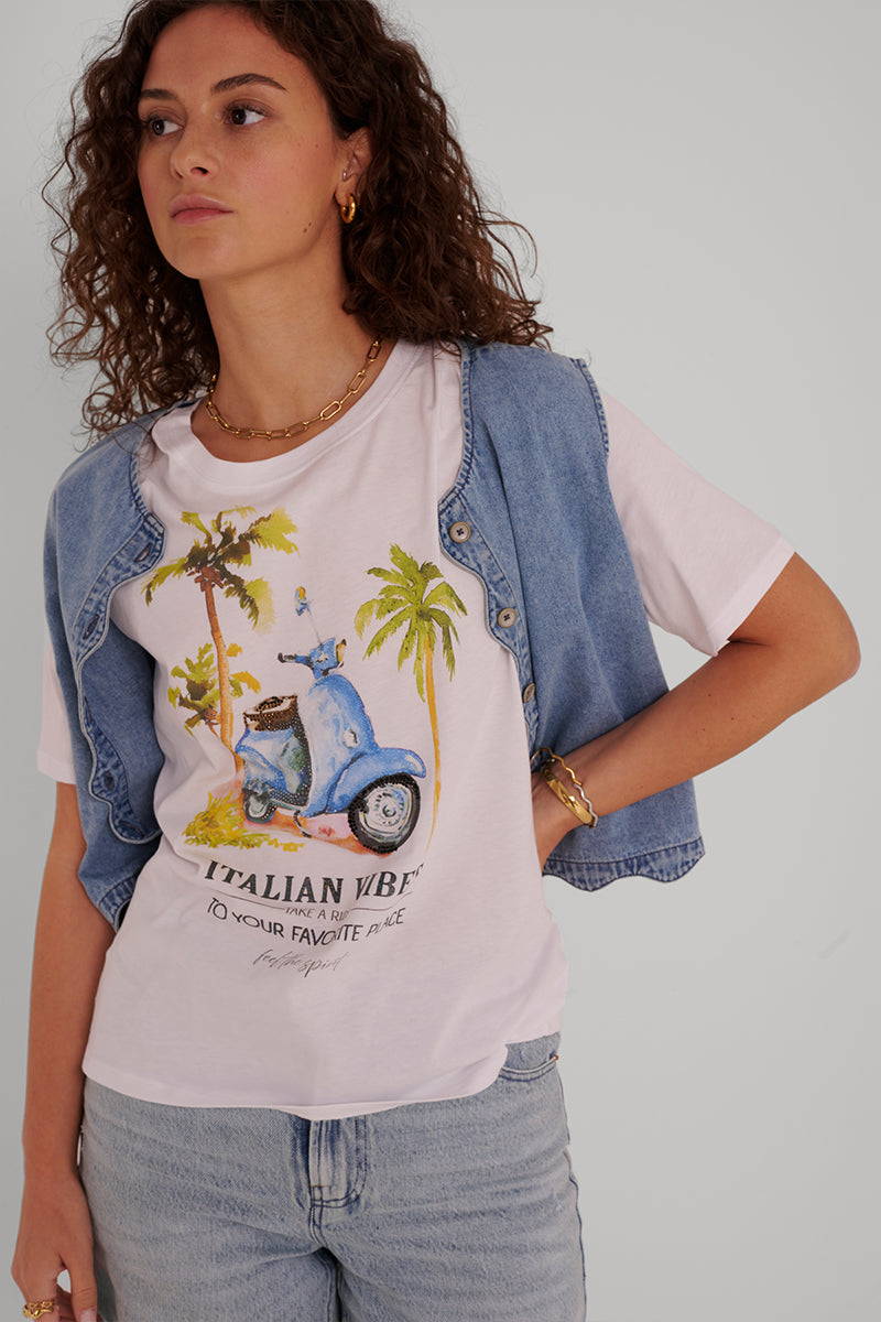 Eine Frau mit lockigen braunen Haaren trägt ein weißes T-Shirt von Princess Goes Hollywood aus Baumwolle, auf dem eine blaue Vespa, Palmen und "Italian Vibes, To Your Favorite Place" abgebildet sind. Dazu trägt sie eine helle Jeansweste, Jeans und Goldschmuck vor einem schlichten Hintergrund.