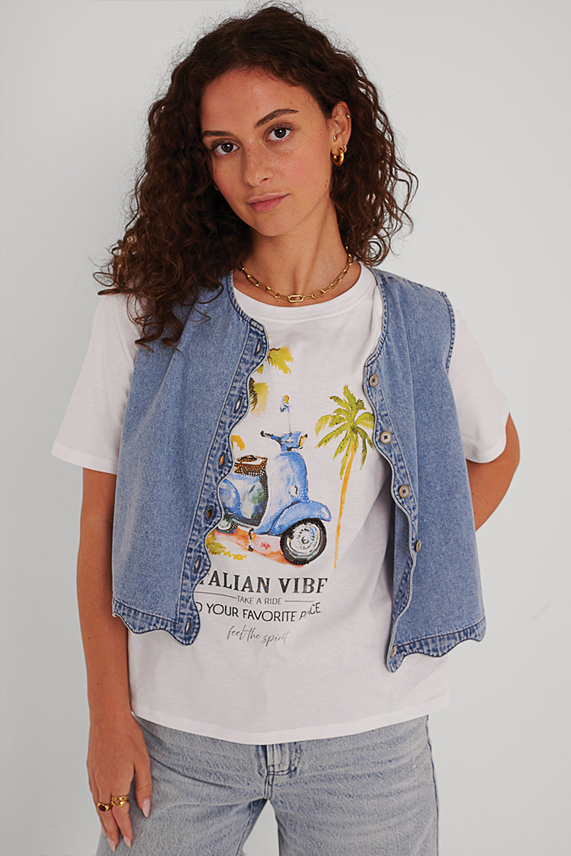 Eine Frau mit lockigem braunem Haar trägt eine ärmellose hellblaue Jeansweste von Princess Goes Hollywood über einem weißen T-Shirt mit einem blauen Motorroller, einer Palme und dem Aufdruck "ITALIAN VIBE". Sie kombiniert es mit hellen Jeans und Goldschmuck vor einem hellen Hintergrund.