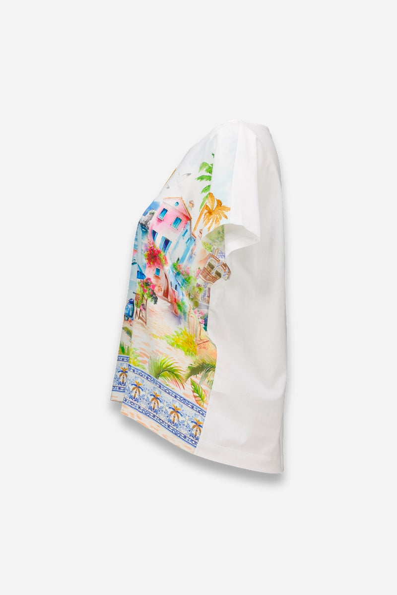 Seitenansicht eines weißen Princess Goes Hollywood Positano-Print T-Shirt aus Baumwolle, mit bunten Pastellhäusern, Grünzeug, Palmen und einer blauen Zierborte entlang des Saums auf einem schlichten weißen Hintergrund.