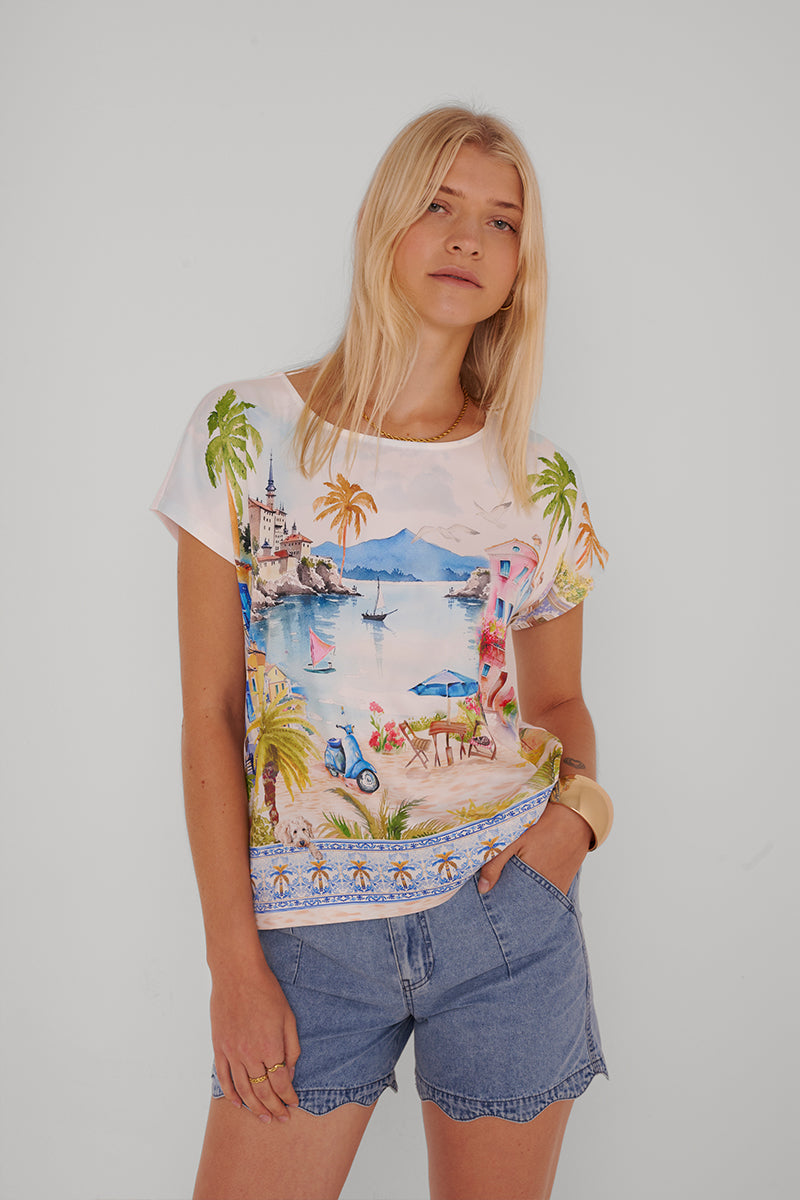 Eine Frau mit langen blonden Haaren trägt eine Princess Goes Hollywood-Jeanshose mit Gummibund und ein buntes T-Shirt mit Meerblick. Sie steht entspannt vor einem schlichten weißen Hintergrund, eine Hand in der Tasche.