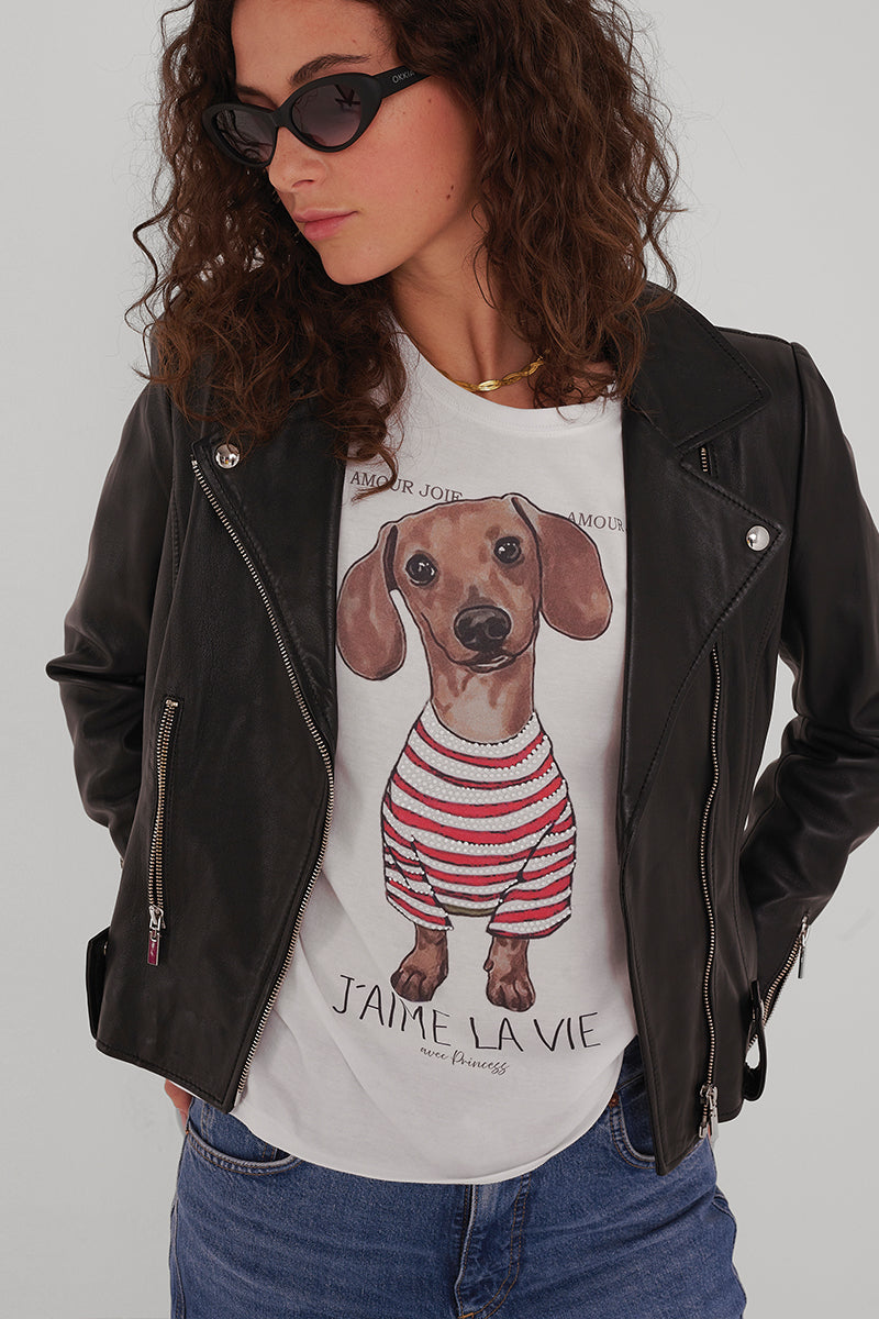 Eine Frau mit lockigem Haar trägt eine schwarze Sonnenbrille, eine schwarze Lederjacke, blaue Jeans und ein Princess Goes Hollywood Baumwolle T-Shirt mit einem niedlichen Dackel-Print eines Hundes in einem rot-gestreiften Hemd und französischem Text, der vor einem schlichten Hintergrund steht.