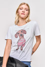 Eine blonde Frau trägt ein Princess Goes Hollywood T-Shirt aus 100% Baumwolle mit Dackel-Aufdruck, roten und weißen Streifen und französischem Text. Sie steht vor einem einfarbigen Hintergrund und hält eine schwarze Tasche mit einem Arm hinter ihrem Rücken.