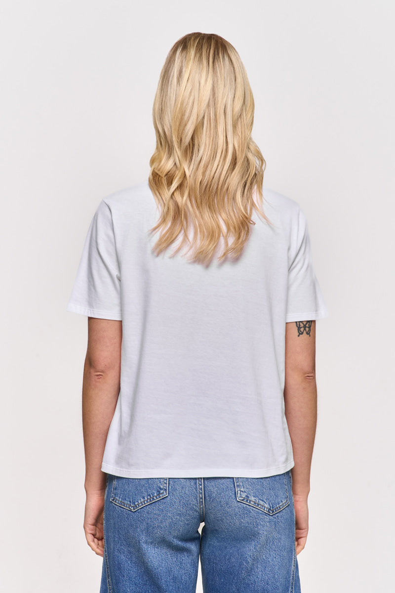Eine Person mit langen, gewellten blonden Haaren ist von hinten zu sehen. Sie trägt ein schlichtes weißes Princess Goes Hollywood Baumwolle T-Shirt und blaue Jeans. Der Hintergrund ist weiß, und auf dem linken Oberarm befindet sich eine kleine Tätowierung.