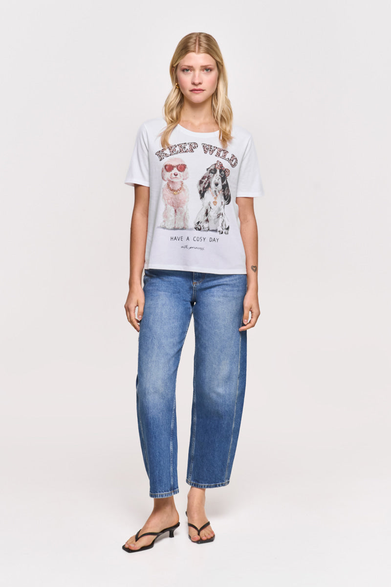 Eine Frau mit glattem blondem Haar trägt ein weißes Hunde-Print T-Shirt aus Baumwolle von Princess Goes Hollywood mit zwei Hunde-Illustrationen und dem Spruch "STAY WILD HAVE A COSY DAY", dazu blaue Jeans und schwarze Sandalen.