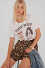 Eine blonde Frau trägt ein weißes "Keep Wild"-T-Shirt von Princess Goes Hollywood mit Hunden auf einer Sonnenbrille und einer Schleife mit Leopardenmuster, dazu hochgeschnittene Leoprint-Shorts. Sie hält eine Jeansjacke in der Hand und trägt ein goldenes Armband, während sie ruhig in die Kamera schaut.