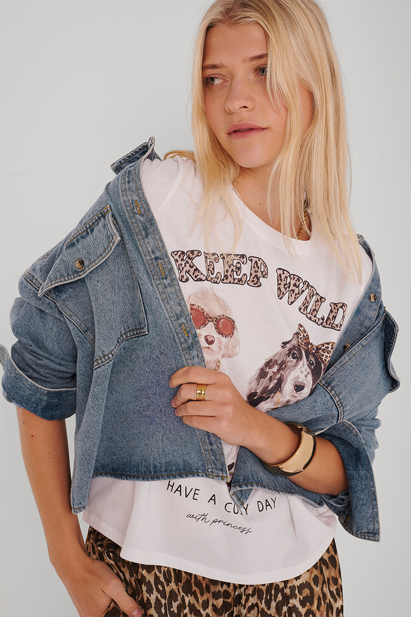 Eine blonde Frau trägt eine Princess Goes Hollywood Damen Jeansjacke über einem weißen grafischen T-Shirt mit Hundeillustrationen und dem Text "KEEP WILD", gepaart mit einer Hose mit Leopardenmuster. Sie ist leicht geschminkt, trägt goldene Ringe und blickt leicht zur Seite mit einem neutralen Ausdruck.