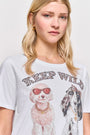 Eine blonde Frau trägt ein Princess Goes Hollywood Keep Wild Shirt, ein Baumwoll-T-Shirt mit einem Aufdruck von zwei Hunden mit Sonnenbrillen und Schmuck und dem fetten Schriftzug "KEEP WILD" auf einem schlichten hellen Hintergrund.