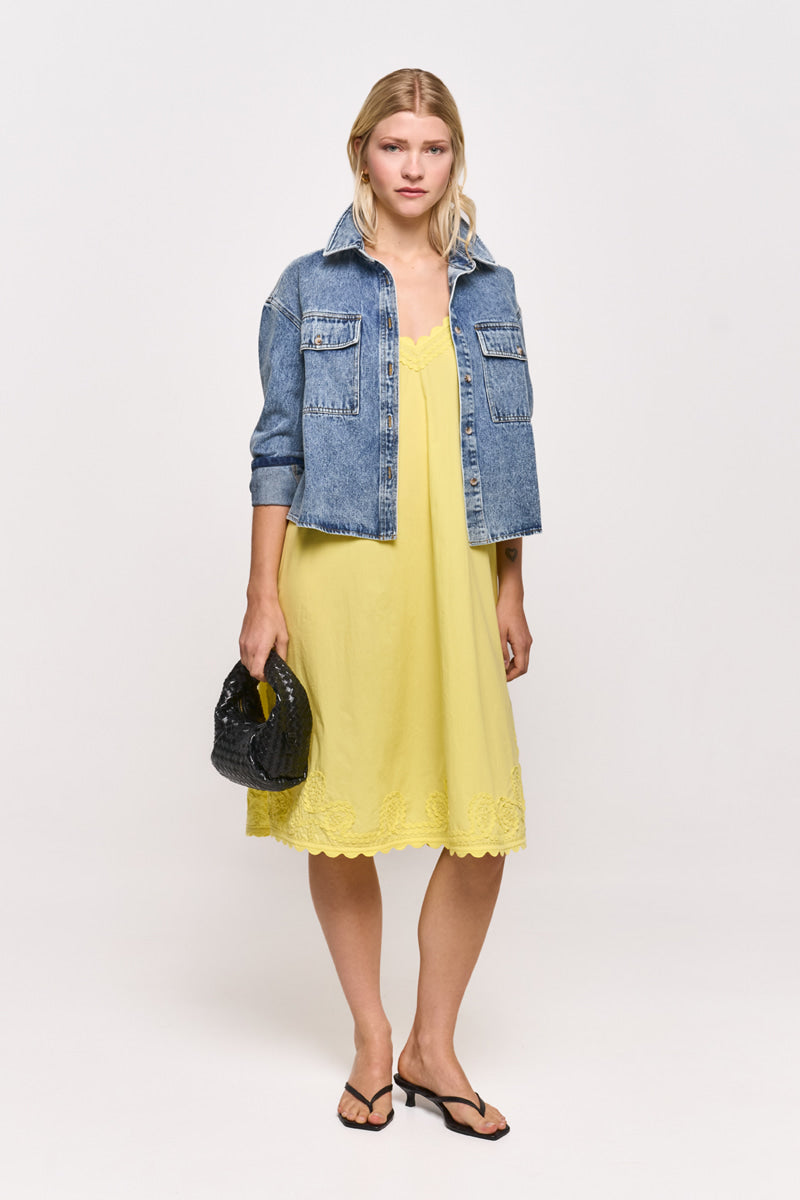 Eine Frau mit blondem, mittellangem Haar trägt ein lockeres gelbes Midikleid von Princess Goes Hollywood mit Stickereien und Spitzenbesatz, eine offene blaue Jeansjacke, schwarze Flip-Flop-Sandalen und eine kleine schwarze, strukturierte Handtasche vor einem schlichten Hintergrund.