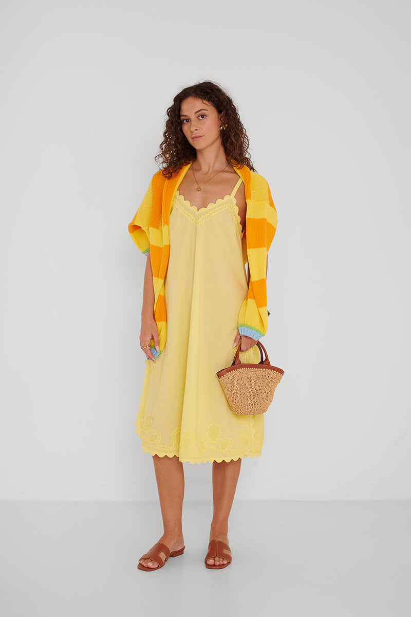Eine Frau mit lockigem braunem Haar steht vor einem weißen Hintergrund und trägt ein lockeres, gelbes Baumwoll-Mididress mit Spitzenbesatz von Princess Goes Hollywood, einen gelb-orange karierten Pullover über den Schultern, eine kleine Strohtasche und braune Sandalen.
