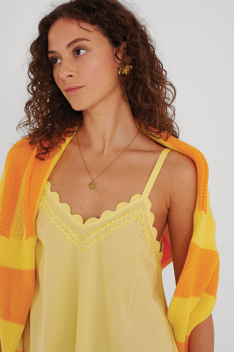 Eine Frau mit lockigem braunem Haar trägt ein hellgelbes Baumwollmieder mit Wellenmuster von Princess Goes Hollywood und eine goldene Halskette. Ein gelb-orange gestreifter Pullover ist über ihre Schultern drapiert. Sie blickt leicht nach links und hat einen ruhigen Gesichtsausdruck vor einem schlichten Hintergrund.