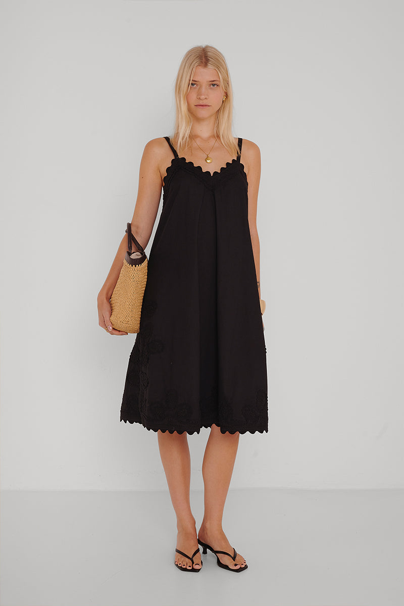 Eine Frau mit langen blonden Haaren steht vor einer weißen Wand und trägt ein schwarzes besticktes Baumwoll-Midikleid mit Wellenschliff von Princess Goes Hollywood. Dazu trägt sie schwarze Sandalen, eine goldene Halskette und eine geflochtene Strohtasche.