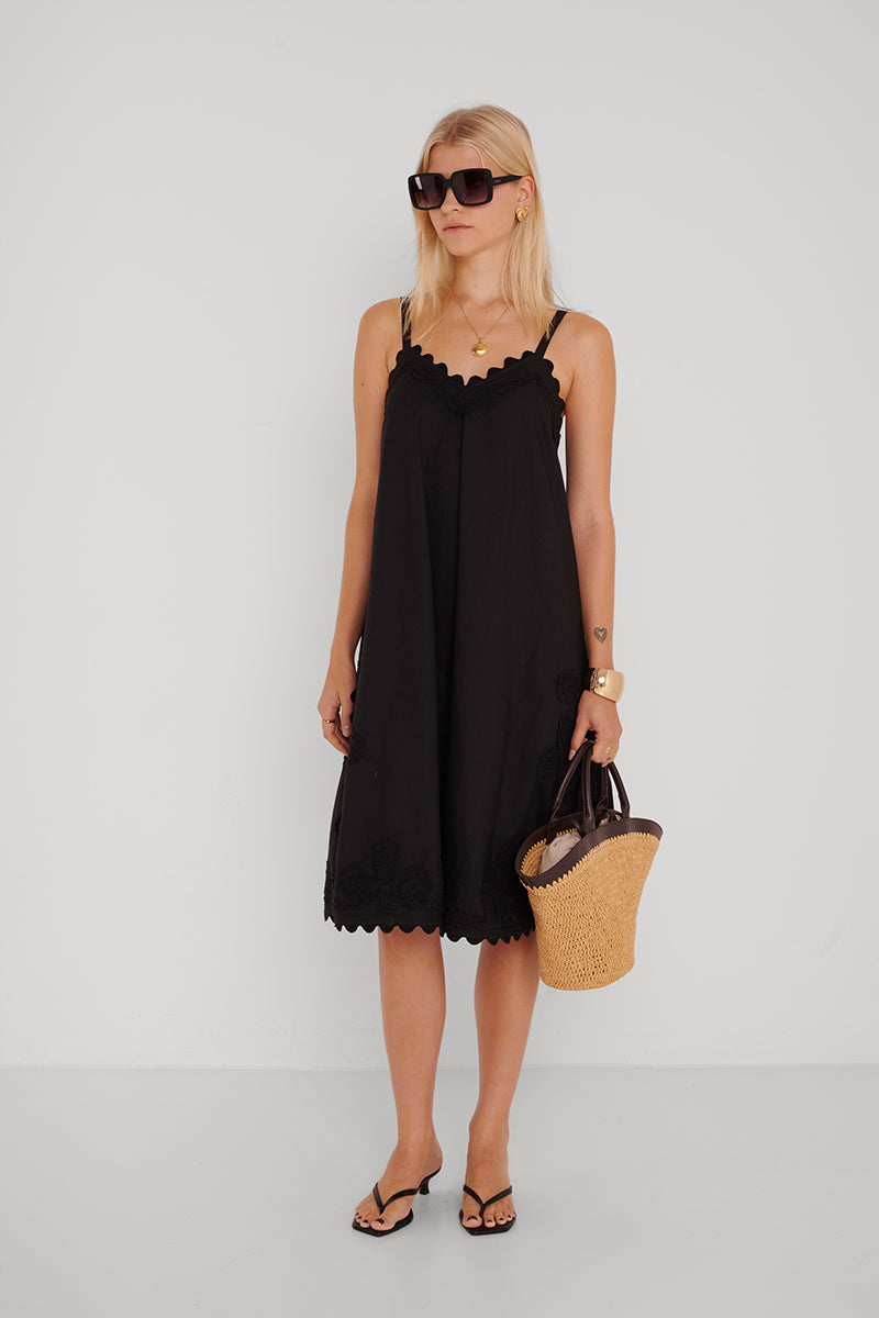 Eine Frau mit blonden Haaren trägt ein schwarzes Midikleid aus Baumwolle mit Stickerei und wellenförmigen Kanten von Princess Goes Hollywood, kombiniert mit großen Sonnenbrillen, goldfarbenem Schmuck, schwarzen Sandalen und einer Strohtasche vor weißem Hintergrund.