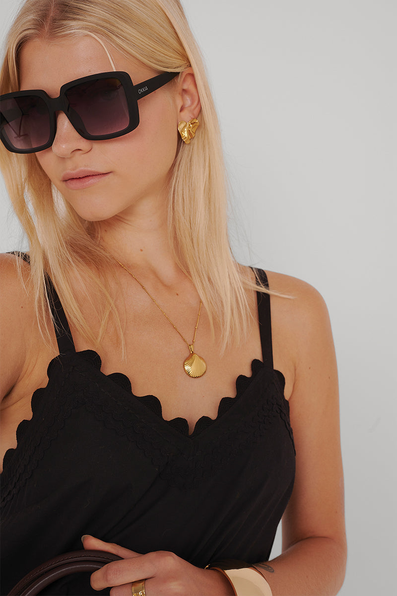 Eine blonde Frau trägt eine übergroße schwarze Sonnenbrille, goldene Muschelohrringe und einen goldenen Anhänger, ein schwarzes Muschel-Camisole unter einem fließenden Princess Goes Hollywood-Midikleid aus Baumwolle, dazu ein goldenes Armband und Ringe.