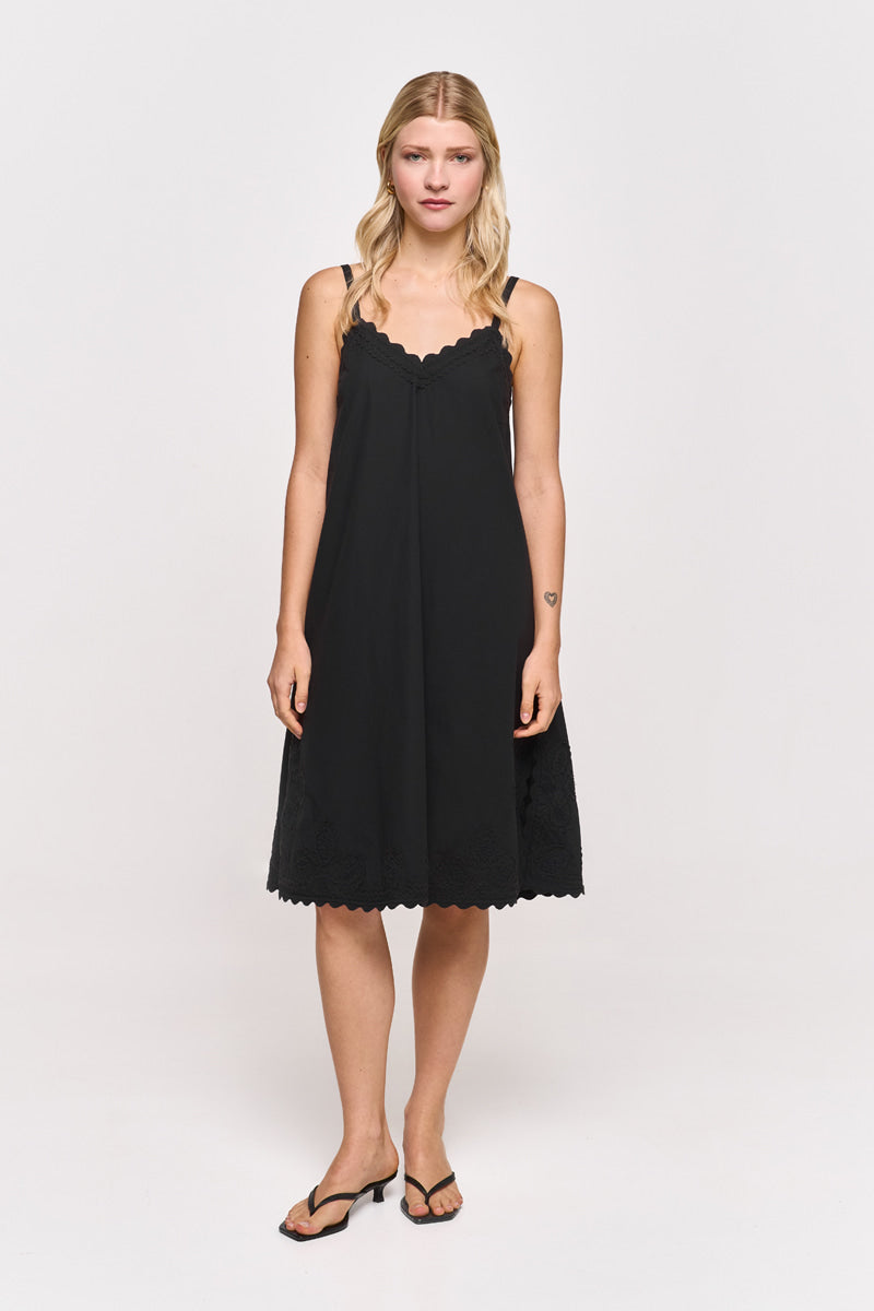 Eine Frau mit langen blonden Haaren steht mit dem Gesicht nach vorne in einem Princess Goes Hollywood Midikleid aus schwarzer Baumwolle mit zarter Stickerei und dünnen Trägern. Sie hat einen neutralen Gesichtsausdruck, trägt schwarze Sandalen und steht vor einem schlichten weißen Hintergrund.