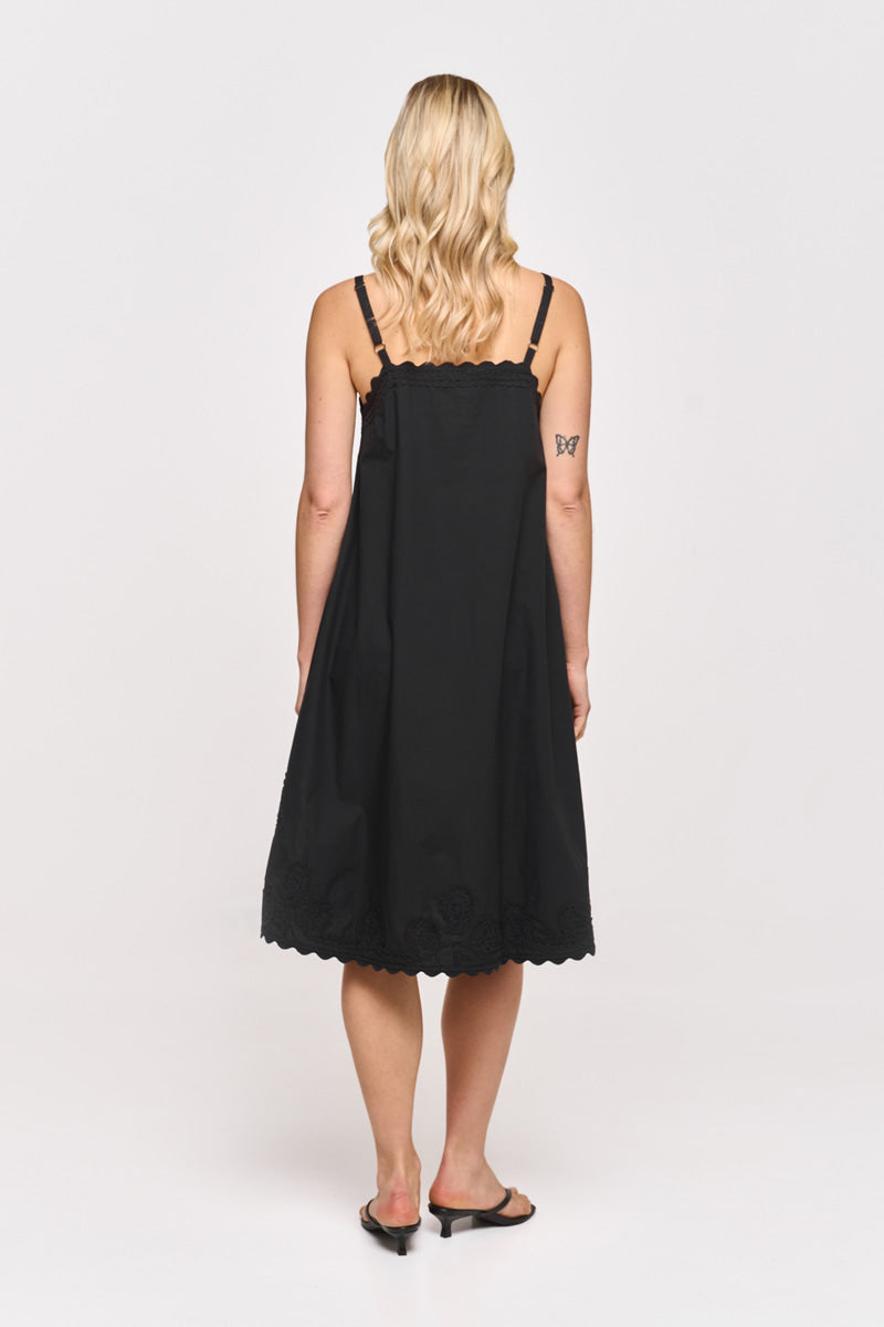Eine Frau mit blondem, gewelltem Haar steht mit dem Rücken zur Kamera in einem ärmellosen, schwarzen Midikleid aus Baumwolle von Princess Goes Hollywood. Sie trägt schwarze hochhackige Sandalen und zeigt ein Schmetterlingstattoo auf ihrem linken Oberarm vor weißem Hintergrund.