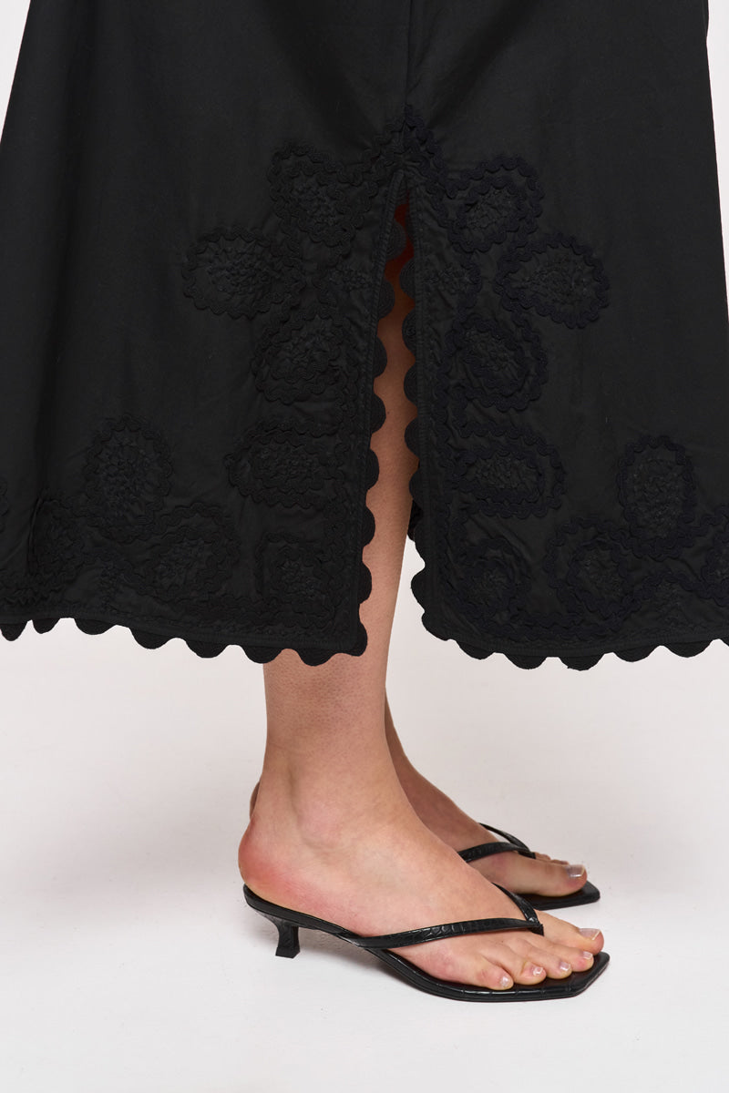 Eine Person trägt einen schwarzen Midirock aus Baumwolle mit floraler Stickerei und wellenförmigen Kanten von Princess Goes Hollywood, dazu schwarze offene Heels. Durch den frontalen Schlitz sind Beine und Füße vor schlichtem Hintergrund sichtbar.