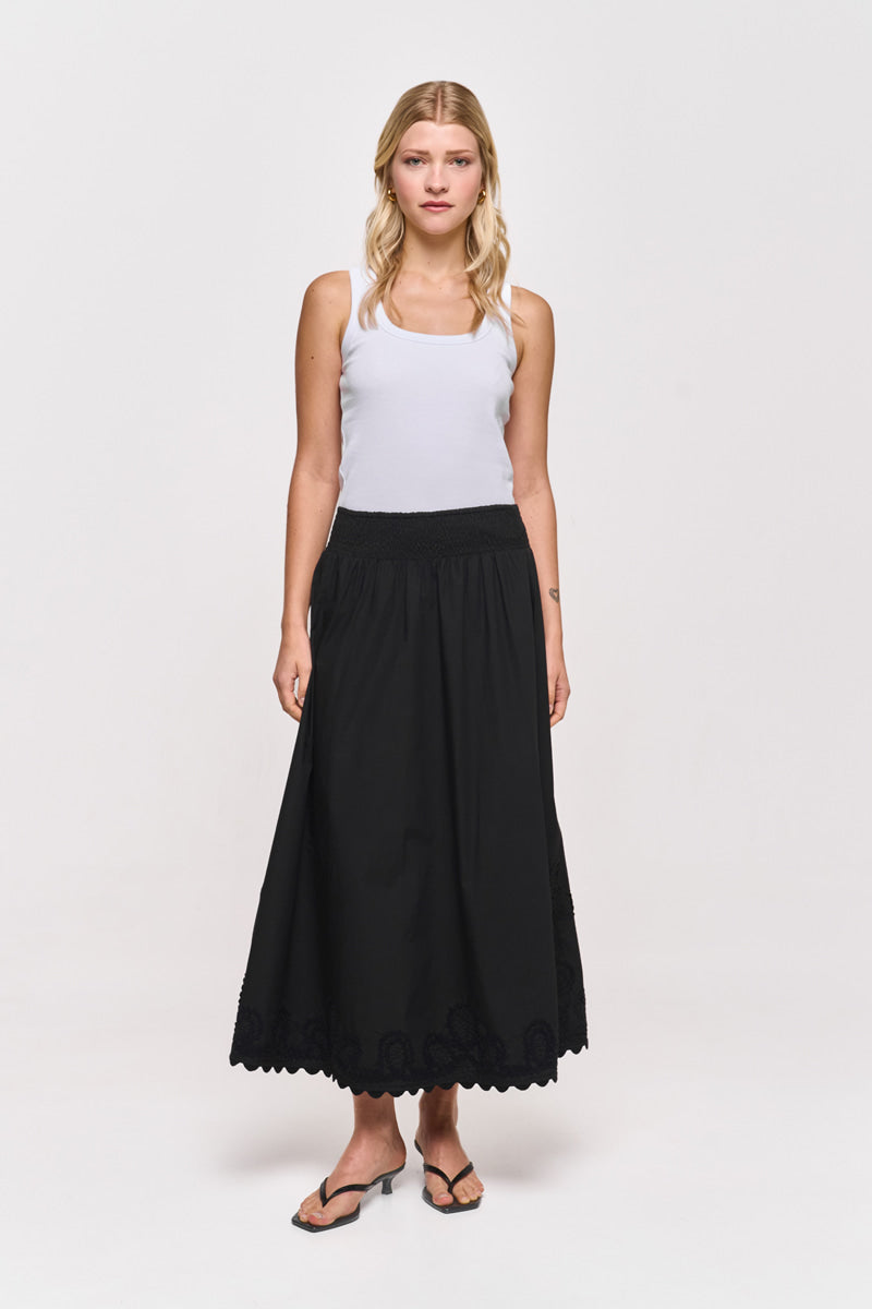 Eine Frau mit langem blondem Haar blickt nach vorne und trägt ein weißes Princess Goes Hollywood Baumwolle-Tanktop, einen langen schwarzen Midirock mit Wellenbesatz und schwarze Flip-Flops. Sie steht vor einem schlichten weißen Hintergrund, ihr Gesichtsausdruck ist neutral.