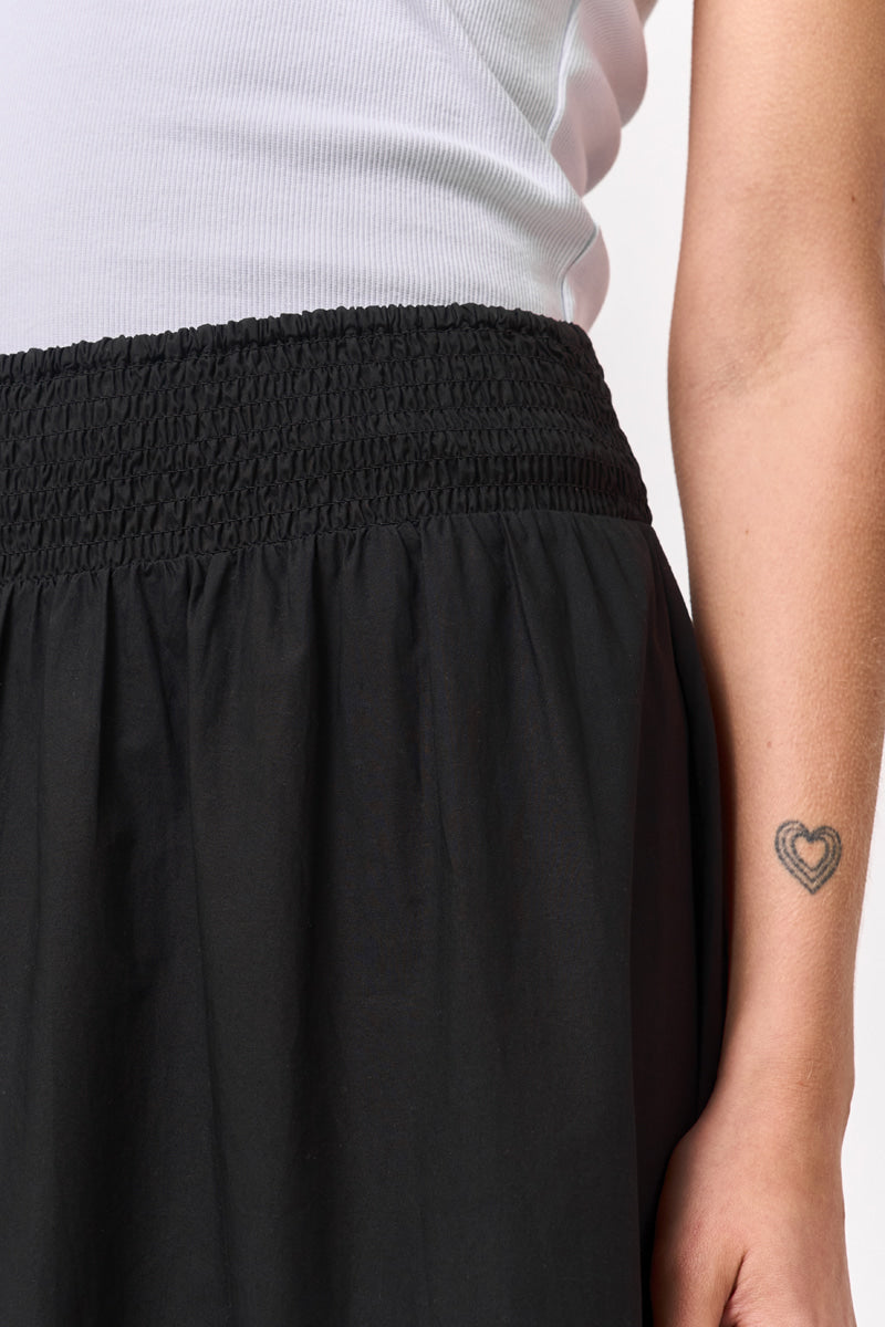 Eine Nahaufnahme zeigt eine Person in einem weißen gerippten Tank-Top und einem schwarzen Princess Goes Hollywood-Midirock mit gerüschtem Gummibund. Auf dem rechten Arm ist ein doppelt umrandetes Herz-Tattoo vor einem schlichten, hellen Hintergrund zu sehen.