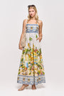 Eine Frau trägt ein ärmelloses Maxikleid von Princess Goes Hollywood aus Baumwolle-Seide-Mix mit Positano-Print, blauen Bordüren und tropischen Palmenprints sowie eine geflochtene Schultertasche, beigefarbene Heels und Sonnenbrille.