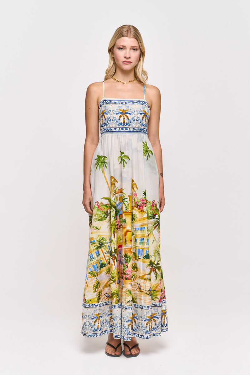 Eine junge Frau blickt nach vorne und trägt ein Princess Goes Hollywood Positano-Print Maxikleid aus Baumwolle-Seide-Mix. Das ärmellose Kleid zeigt lebhafte tropische Szenen mit Palmen und Häusern sowie blau gemusterte Bordüren an Mieder und Saum, dazu Sandalen.