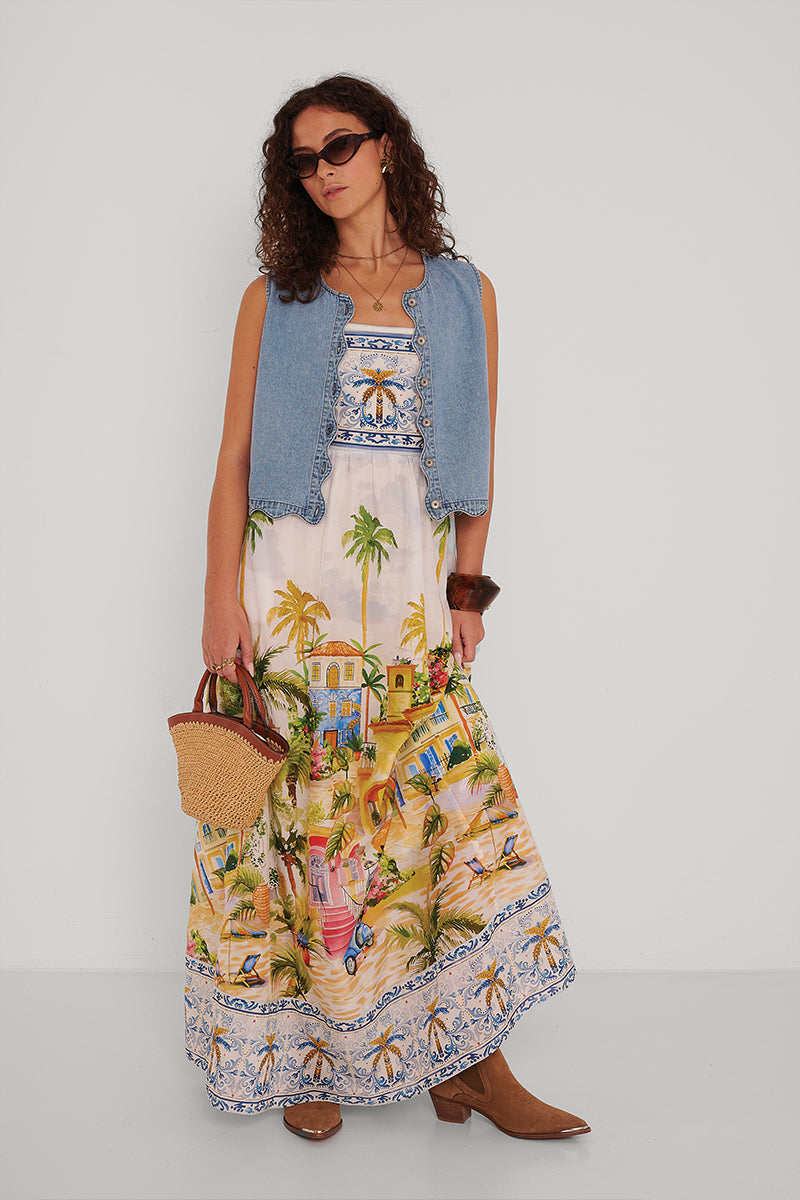 Eine Frau steht vor einer weißen Wand und trägt eine hellblaue, ärmellose Jeansweste von Princess Goes Hollywood über einem bunten Maxikleid mit Positano-Print. Dazu trägt sie eine braune Sonnenbrille, braune Stiefel, einen Armreif, eine Strohhandtasche und lockere, gewellte Haare.