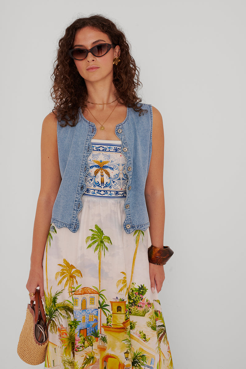 Eine Frau mit lockigem braunem Haar trägt eine Sonnenbrille mit Katzenaugen, eine ärmellose Jeansweste und ein langes Kleid mit einem tropischen Dorfaufdruck. Sie hält eine Strohhandtasche und ein braunes Armband vor einem hellgrauen Hintergrund.