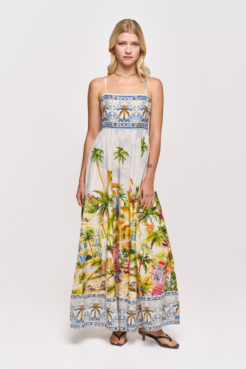 Eine Frau blickt nach vorne und trägt ein ärmelloses, bodenlanges Maxikleid mit Positano-Print von Princess Goes Hollywood aus einem Baumwoll-Seiden-Gemisch. Das Kleid zeigt Palmen und Häuser am Saum, blaue Zierborten und wird mit Sandalen gestylt.
