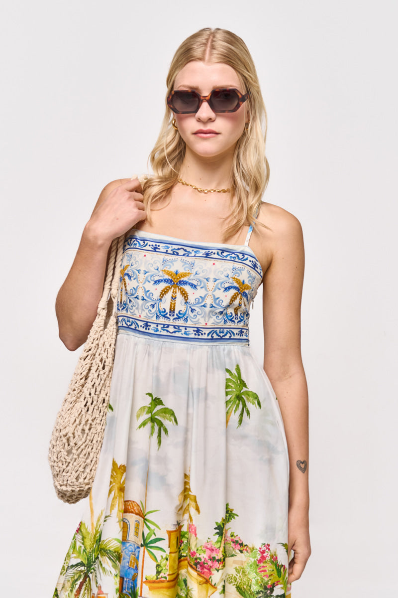 Eine blonde Frau trägt eine große braune Sonnenbrille und ein trägerloses Princess Goes Hollywood Maxikleid mit einem bunten Positano-Aufdruck. Sie hält eine gewebte beigefarbene Tasche in der Hand und zeigt eine kleine Herz-Tätowierung auf ihrem Unterarm, während sie vor einem schlichten hellen Hintergrund steht.