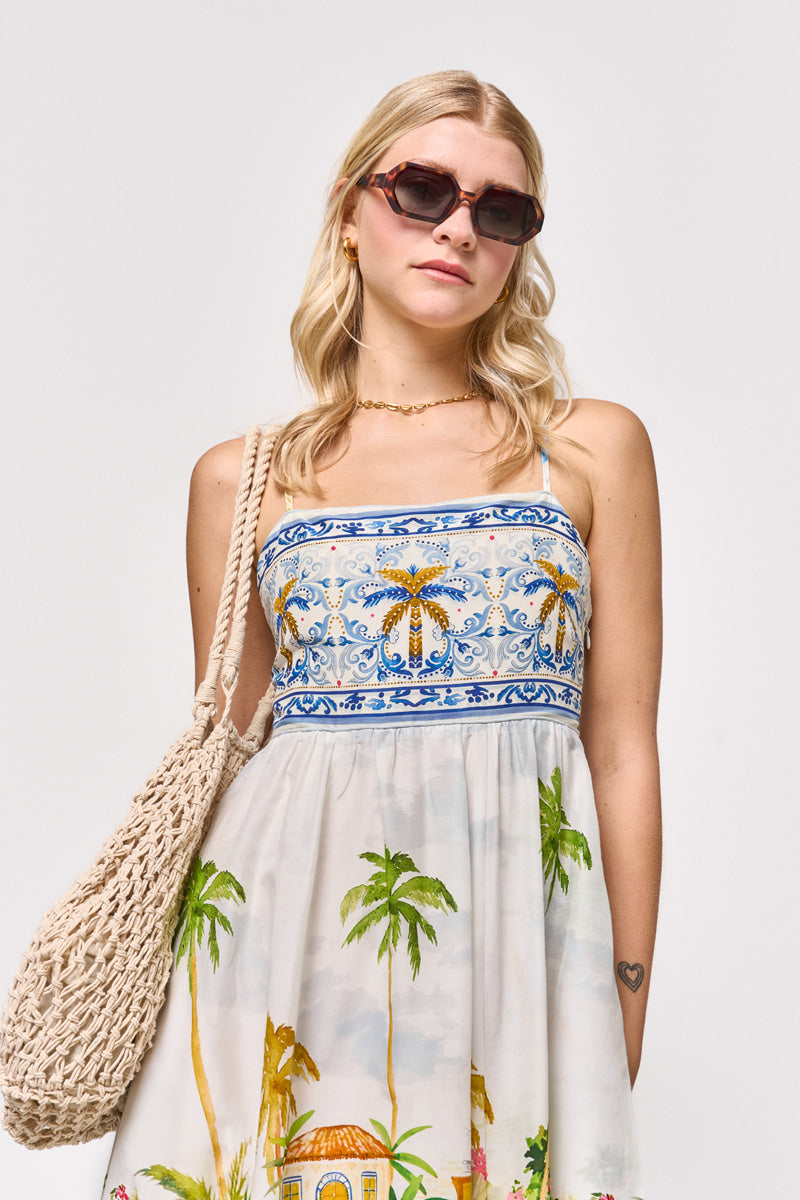 Eine Frau mit langen blonden Haaren trägt eine Sonnenbrille, eine goldene Halskette und ein ärmelloses Princess Goes Hollywood Maxikleid mit Positano-Print aus einer Baumwoll-Seidenmischung. Sie trägt eine gewebte Umhängetasche und zeigt ein kleines Herz-Tattoo auf ihrem linken Unterarm.