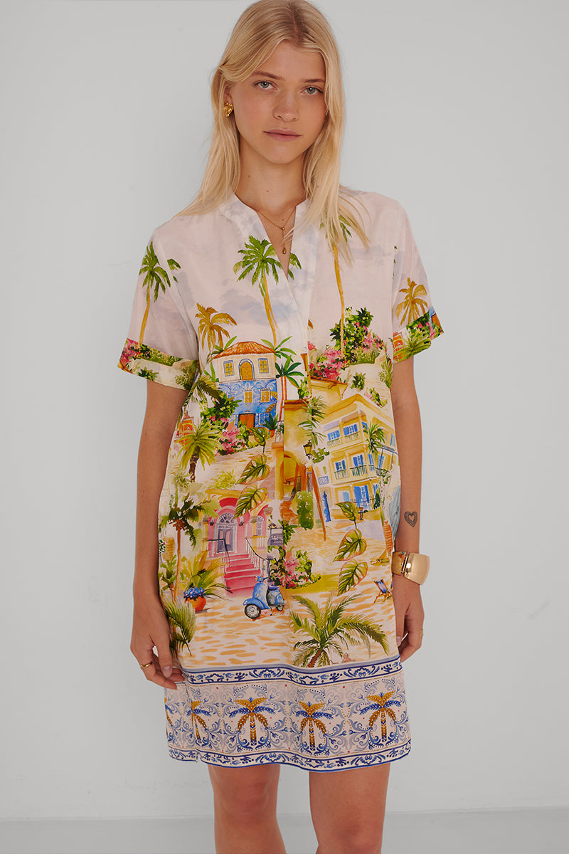 Eine Frau mit glattem blondem Haar trägt ein Princess Goes Hollywood Blusenkleid mit Positano-Print aus einer Baumwoll-Seiden-Mischung, auf dem tropische Häuser, Palmen, Blumen und eine blau-weiße Bordüre am Saum abgebildet sind, während sie vor einer schlichten weißen Wand steht.