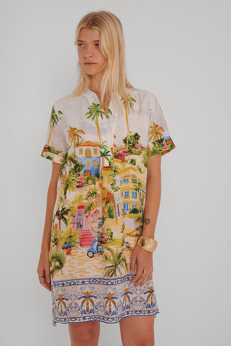 Eine Frau mit langen blonden Haaren trägt ein farbenfrohes Kleid aus Baumwoll-Seide mit Positano-Print, auf dem Palmen, Gebäude, ein blauer Motorroller und ein blau-weiß gemusterter Saum zu sehen sind. Sie steht vor einem schlichten weißen Hintergrund und blickt zur Seite.