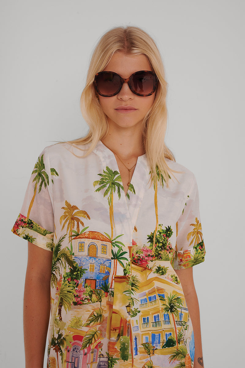 Eine Frau mit langen blonden Haaren trägt eine große braune Sonnenbrille und ein lebhaftes kurzärmeliges Hemd mit Princess Goes Hollywood Positano-Print, auf dem ein tropisches Haus und eine Palme abgebildet sind. Sie steht vor einem schlichten weißen Hintergrund und hat einen neutralen Gesichtsausdruck.