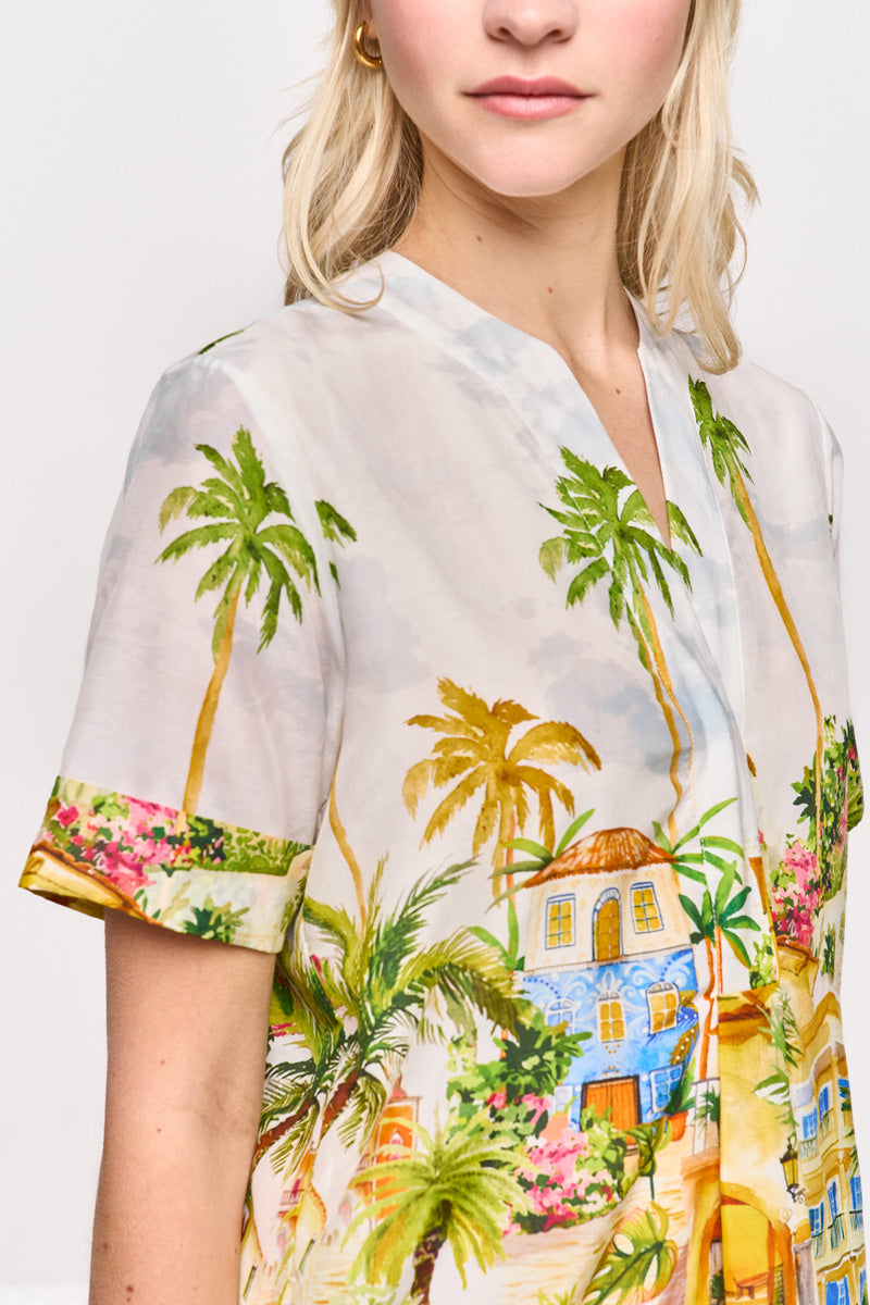 Eine Frau mit heller Haut und blondem Haar modelliert eine Princess Goes Hollywood Bluse mit Positano-Print (Baumwolle-Seide-Mix) mit Palmen, Blumen und pastellfarbenen Gebäuden. Nur ihr unteres Gesicht und ihr Oberkörper sind vor einem hellen Hintergrund zu sehen.