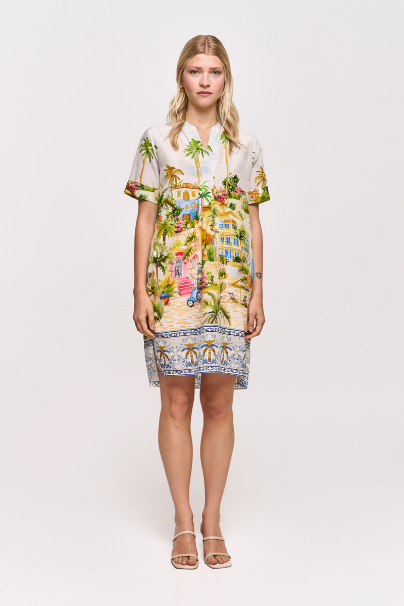 Eine Frau mit glattem blondem Haar trägt ein Princess Goes Hollywood Positano-Print Kleid aus Baumwoll-Seidengemisch mit Palmen, Häusern und Stränden, gepaart mit beigen offenen Sandalen, vor einem schlichten weißen Hintergrund.