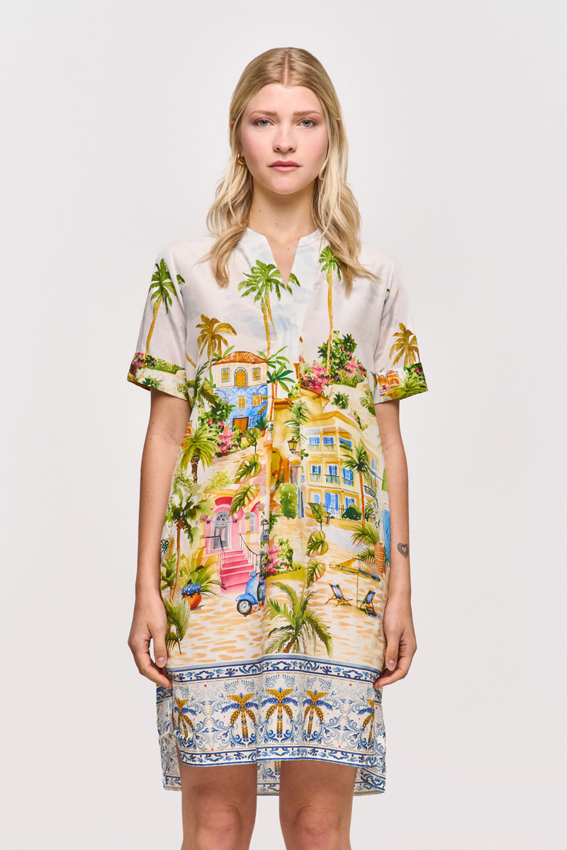 Eine Frau mit glattem blondem Haar blickt nach vorne in einem kurzärmeligen Princess Goes Hollywood Positano-Print Blusenkleid aus einem Baumwolle-Seide-Gemisch mit Palmen, Häusern und einer blauen Bordüre am Saum. Der Hintergrund ist einfarbig weiß.