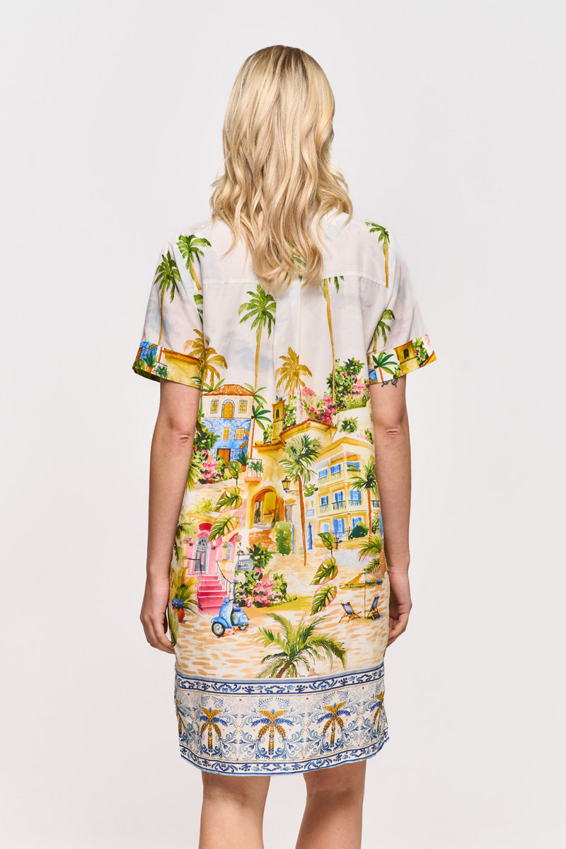 Eine Frau mit gewelltem blondem Haar steht mit dem Gesicht nach unten und trägt ein kurzärmeliges Princess Goes Hollywood Blusenkleid aus Baumwoll-Seiden-Mix mit Positano-Print, mediterranen Palmen, Gebäuden und blauen Mustern an Saum und Ärmeln.