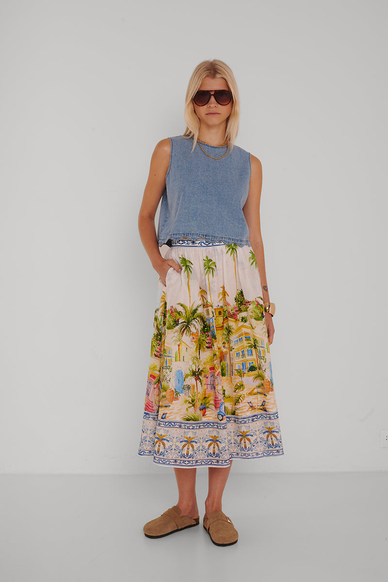 Eine Frau mit kurzen blonden Haaren trägt eine große Sonnenbrille, ein ärmelloses blaues Jeans-Top und einen farbenfrohen Princess Goes Hollywood-Positano-Print-Midirock in üppigem Baumwolle-Seide-Mix. Sie steht vor einer weißen Wand, die Hände in den Rocktaschen, in hellbraunen Slippern.