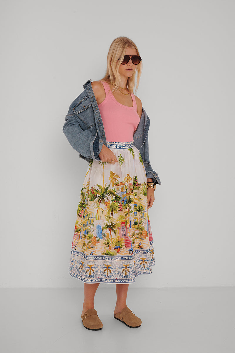 Eine Frau mit blondem Haar steht mit Sonnenbrille vor einer weißen Wand und trägt ein rosa Princess Goes Hollywood Basic Top mit Rundhalsausschnitt aus Baumwolle, eine blaue Jeansjacke über den Schultern, einen Midirock mit tropischem Muster und hellbraune Slipper.