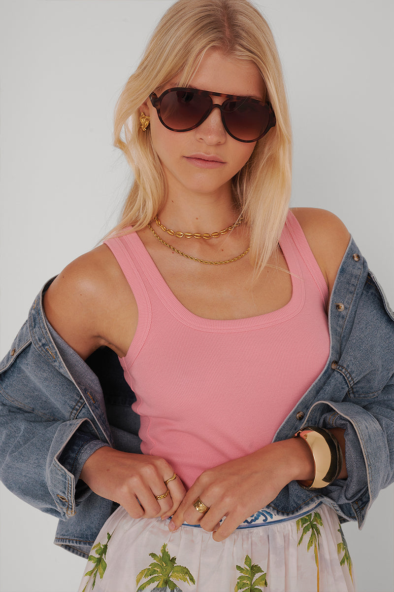 Eine junge Frau mit langen blonden Haaren trägt eine dunkle Sonnenbrille, ein rosafarbenes Princess Goes Hollywood Basic Top mit Rundhalsausschnitt, mehrlagige goldene Halsketten, ein goldenes Armband, eine schulterfreie Jeansjacke und einen Rock mit Palmenmuster vor einem hellen Hintergrund.