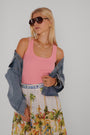 Eine blonde Frau trägt eine große dunkle Sonnenbrille, Goldschmuck und ein rosa Princess Goes Hollywood Basic Top mit Rundhalsausschnitt unter einer drapierten Jeansjacke, gepaart mit einem weißen Rock mit einem farbenfrohen tropischen City-Print vor einem schlichten hellen Hintergrund.