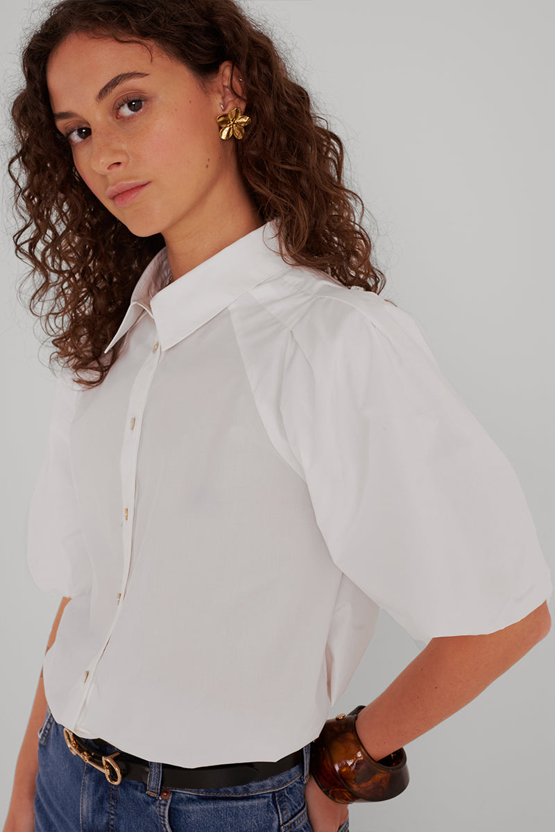 Eine Frau mit lockigem braunem Haar trägt eine Princess Goes Hollywood weiße Baumwollbluse mit 3/4-Puffärmeln, Blue Jeans, einen braunen Armreif und einen goldenen Blumenohrring. Sie steht seitlich und schaut in die Kamera vor einem hellen Hintergrund.
