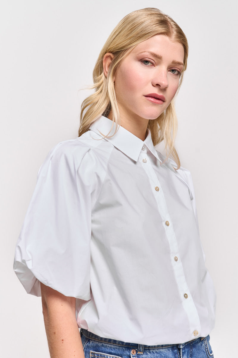 Eine Frau mit langem blondem Haar modelliert eine Princess Goes Hollywood weiße Baumwollbluse mit Hemdkragen und hellbraunen Knöpfen. Sie steht vor einem schlichten, hellen Hintergrund und blickt seitlich mit neutralem Gesichtsausdruck.