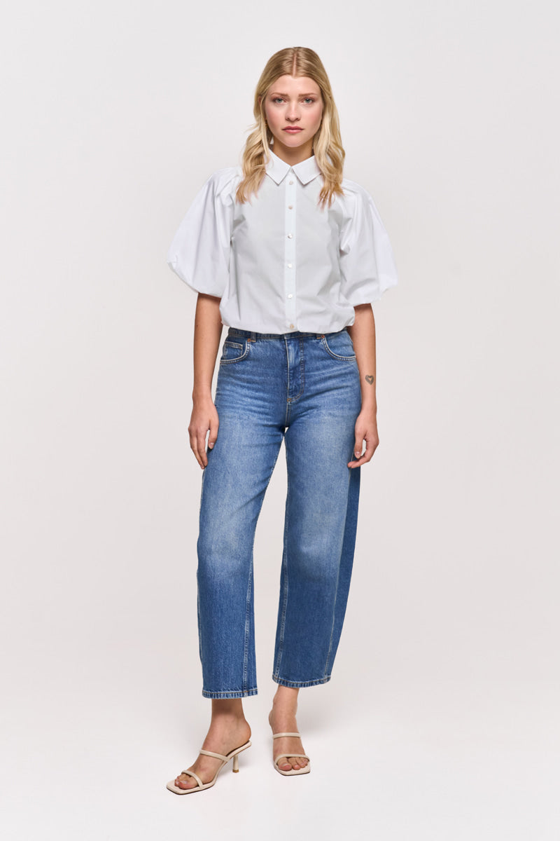 Eine Frau mit glattem, schulterlangem blondem Haar trägt eine Princess Goes Hollywood weiße Baumwollbluse mit Hemdkragen und 3/4-Puffärmel, blaue High-Waisted-Jeans und beigefarbene Sandaletten mit Absatz vor einem schlichten weißen Hintergrund.