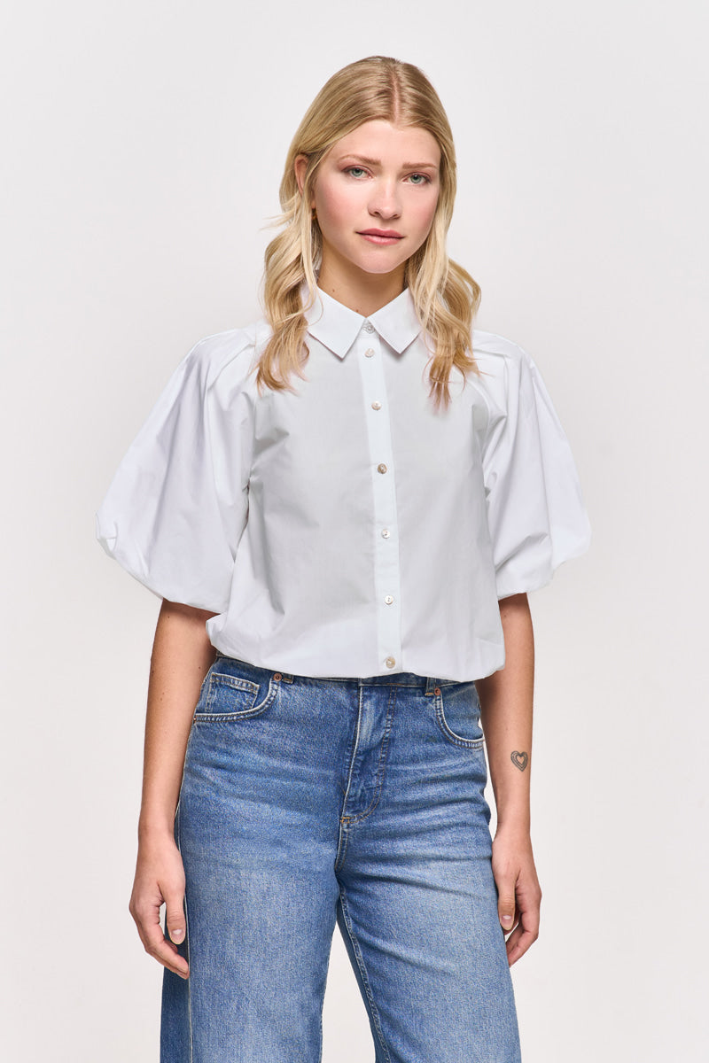 Eine junge Frau mit langen blonden Haaren trägt eine weiße Baumwollbluse von Princess Goes Hollywood mit Hemdkragen und 3/4-Puffärmel zu blauen Jeans. Sie steht frontal vor weißem Hintergrund, Hände an den Seiten, ein kleines Herz-Tattoo am linken Unterarm.