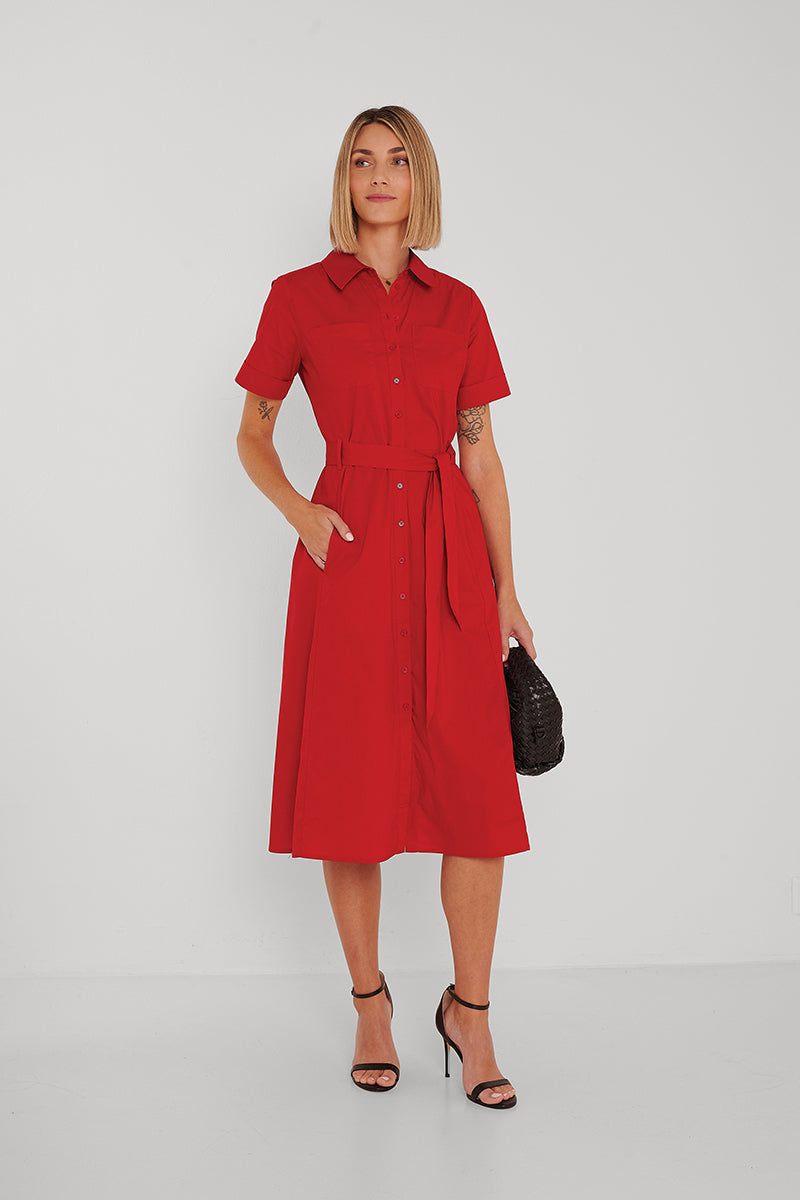 Eine Frau mit Bob trägt ein rotes Midikleid aus Baumwollpopeline mit Knopfleiste und Taillengürtel von Princess Goes Hollywood. Sie hat Arm-Tattoos, hält eine schwarze Clutch und trägt schwarze High Heels vor weißem Hintergrund.